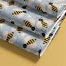 Contact paper 1947: Contact paper with a playful pattern of yellow and black striped bees with beige wings buzzing over white daisy illustrations on a soft slate blue background, displayed folded over a golden yellow surface.