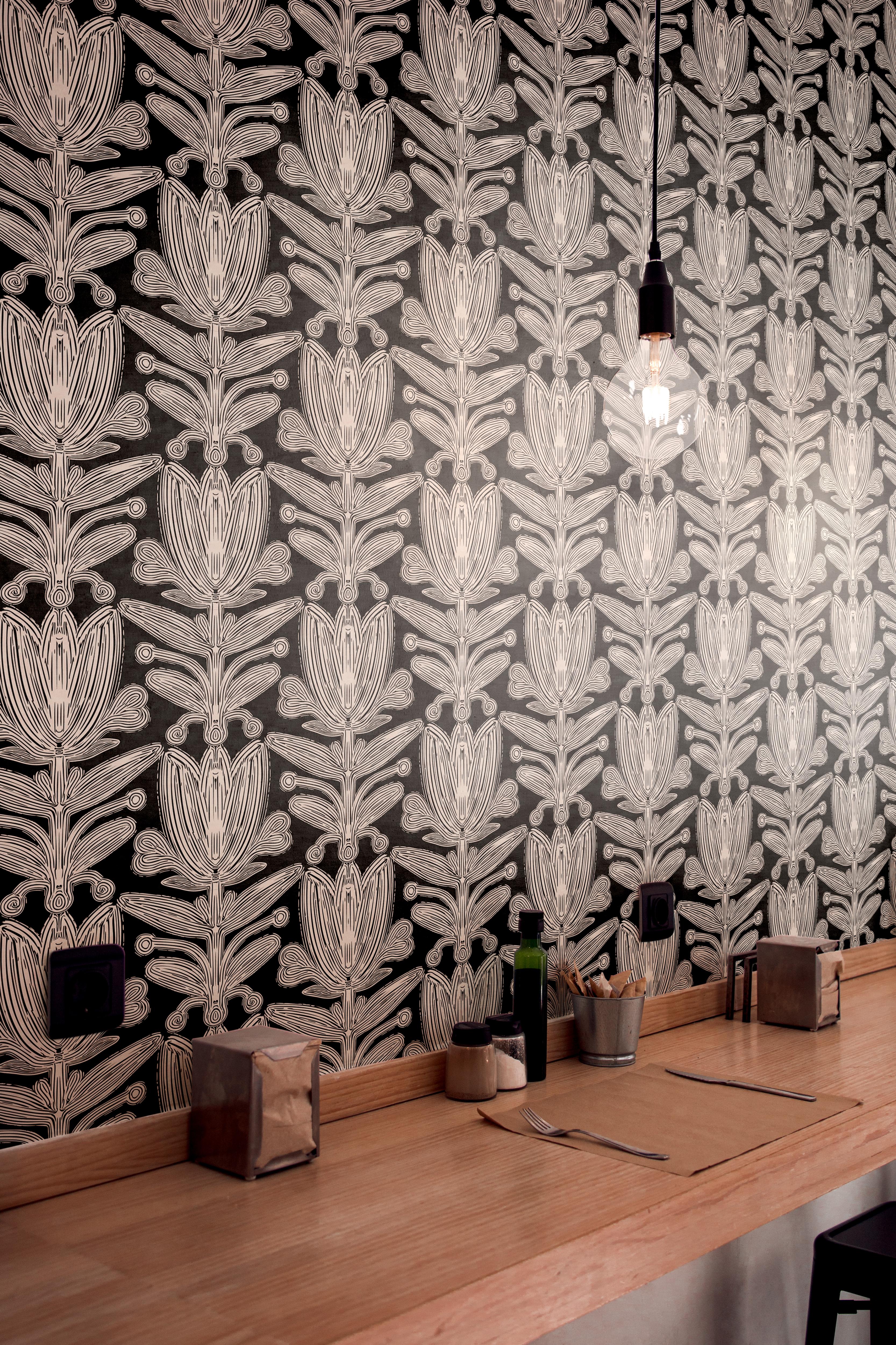 Wallpaper 1960: Black wallpaper with bold, abstract floral line art in vertical columns, creating a dramatic symmetrical look behind a light wood cafe counter.