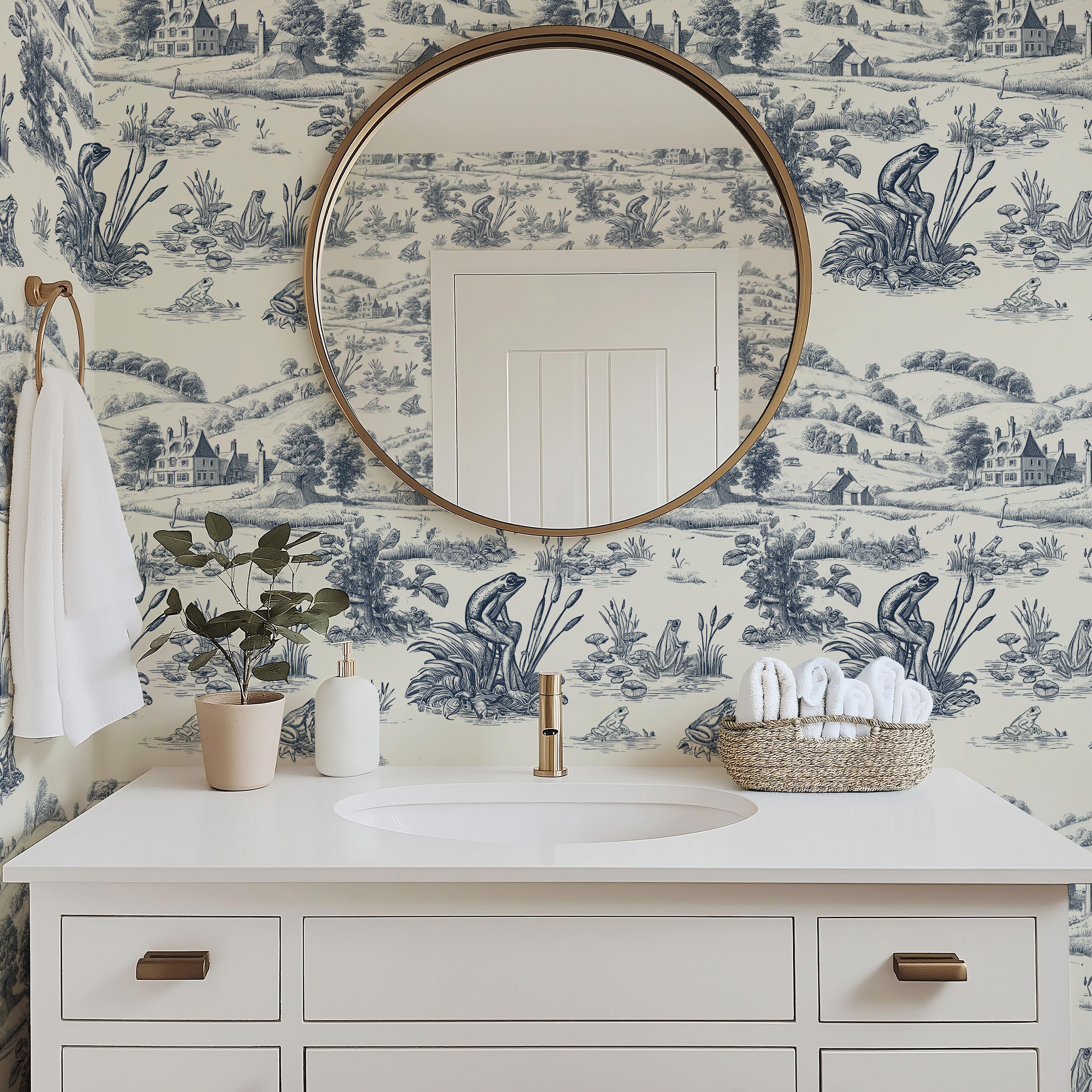 Wallpaper 1961: White bathroom vanity with round gold mirror against a scenic blue and white frog toile wallpaper featuring countryside villages and nature scenes.

