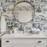 Wallpaper 1961: White bathroom vanity with round gold mirror against a scenic blue and white frog toile wallpaper featuring countryside villages and nature scenes.


