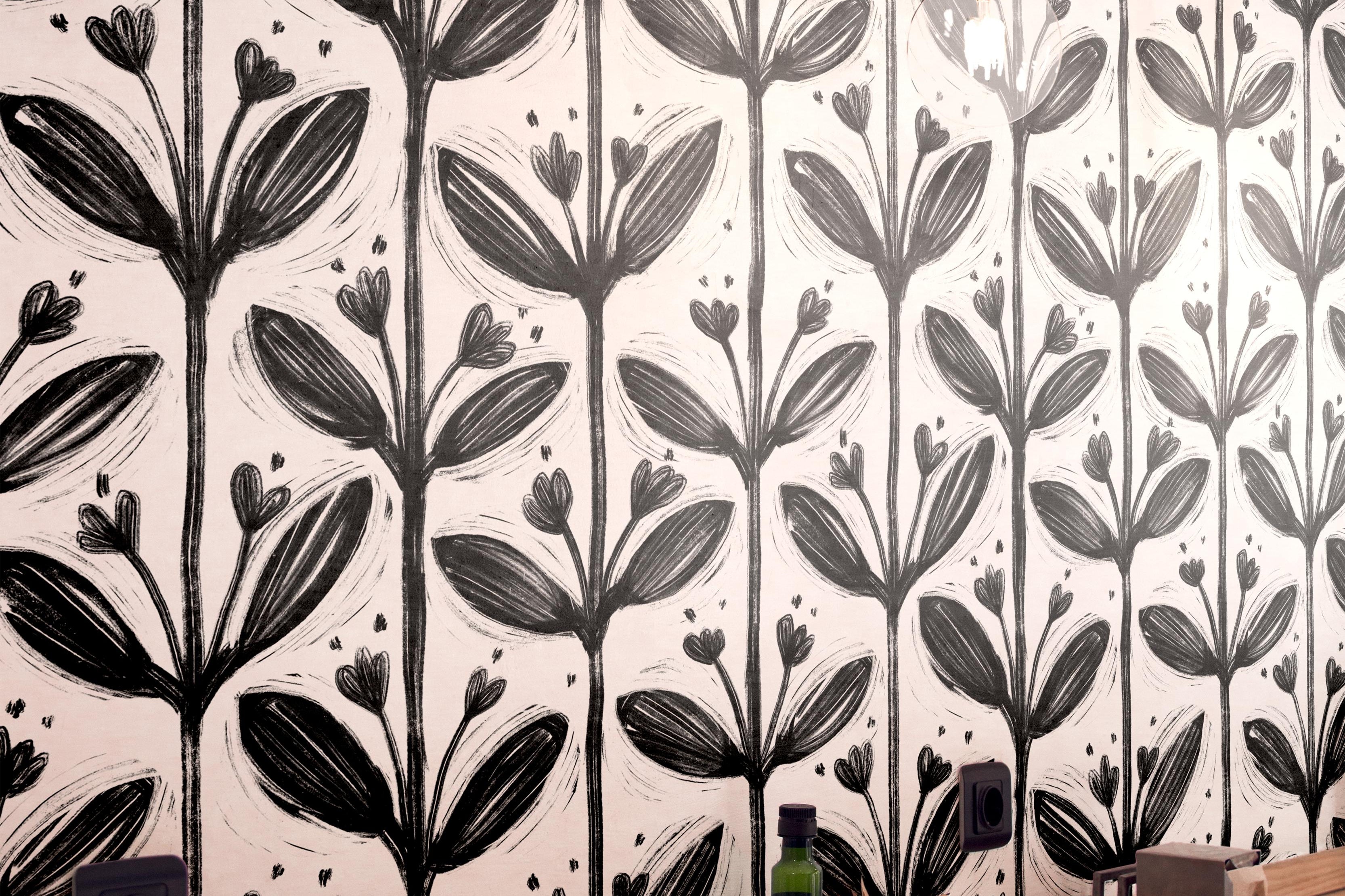 Wallpaper 1982: Modern café interior with black and white bold floral wallpaper featuring large brushstroke flowers, behind a light wood countertop with simple dining setup.

