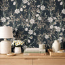 Dark teal floral wallpaper with soft ivory wildflowers and muted green foliage styled above a natural wood dresser with neutral ceramic decor.
