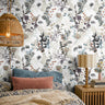 Ivory botanical wallpaper with delicate wildflowers and leafy stems in charcoal, muted blue, sage, taupe, and soft orange, styled in a cozy bedroom with natural wood and layered neutral bedding.