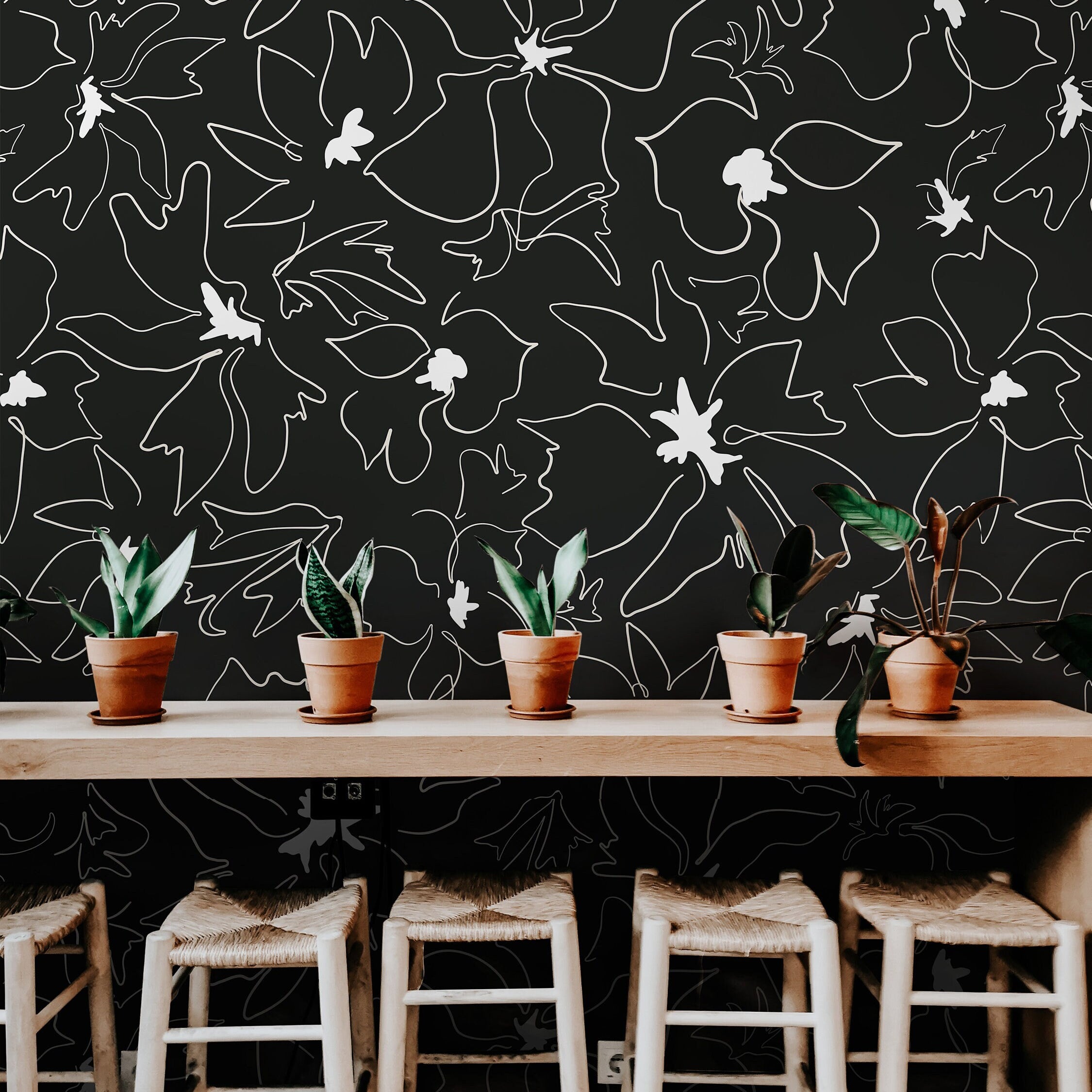 Wallpaper 1158a: Black wallpaper featuring abstract white line-drawn flowers behind a modern wood counter with potted plants and woven stools.

