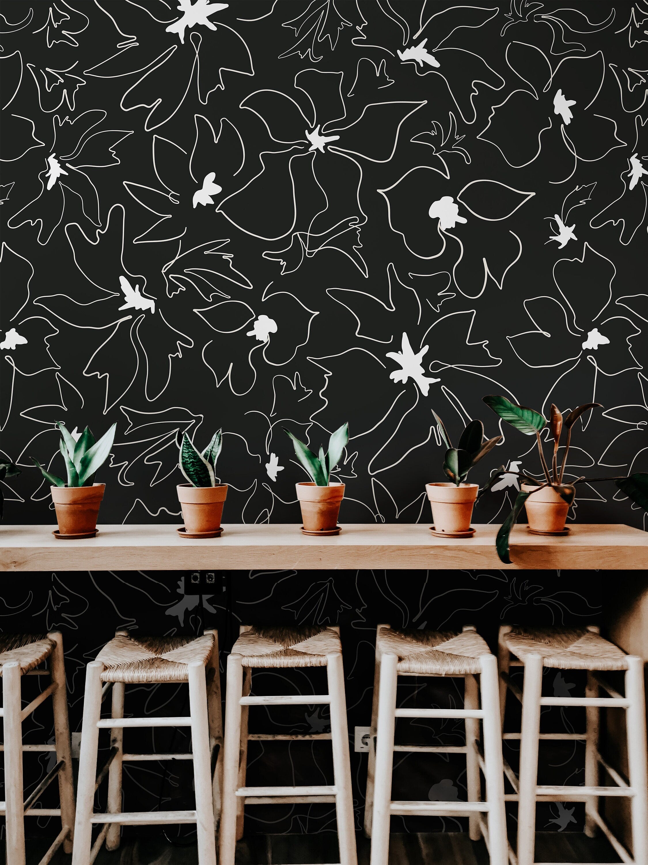 Wallpaper 1158a: Black wallpaper featuring abstract white line-drawn flowers behind a modern wood counter with potted plants and woven stools.

