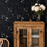 ink and elderflower 3552 moody floral wallpaper behind a rustic wood cabinet with white flowers in a vase and a wood chair