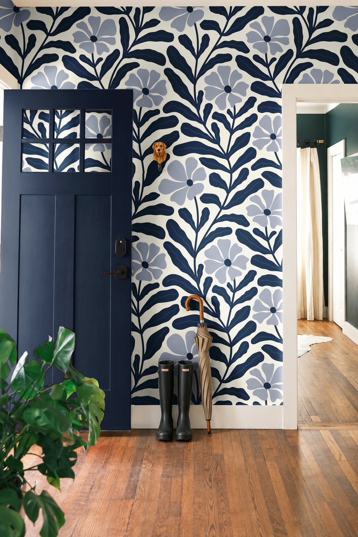 Navy Nature Floral 540 wallpaper featuring oversized dusty blue flowers and dark navy botanical leaves on a cream background, styled in a modern entryway with a navy blue door, natural wood floors, and black rain boots.