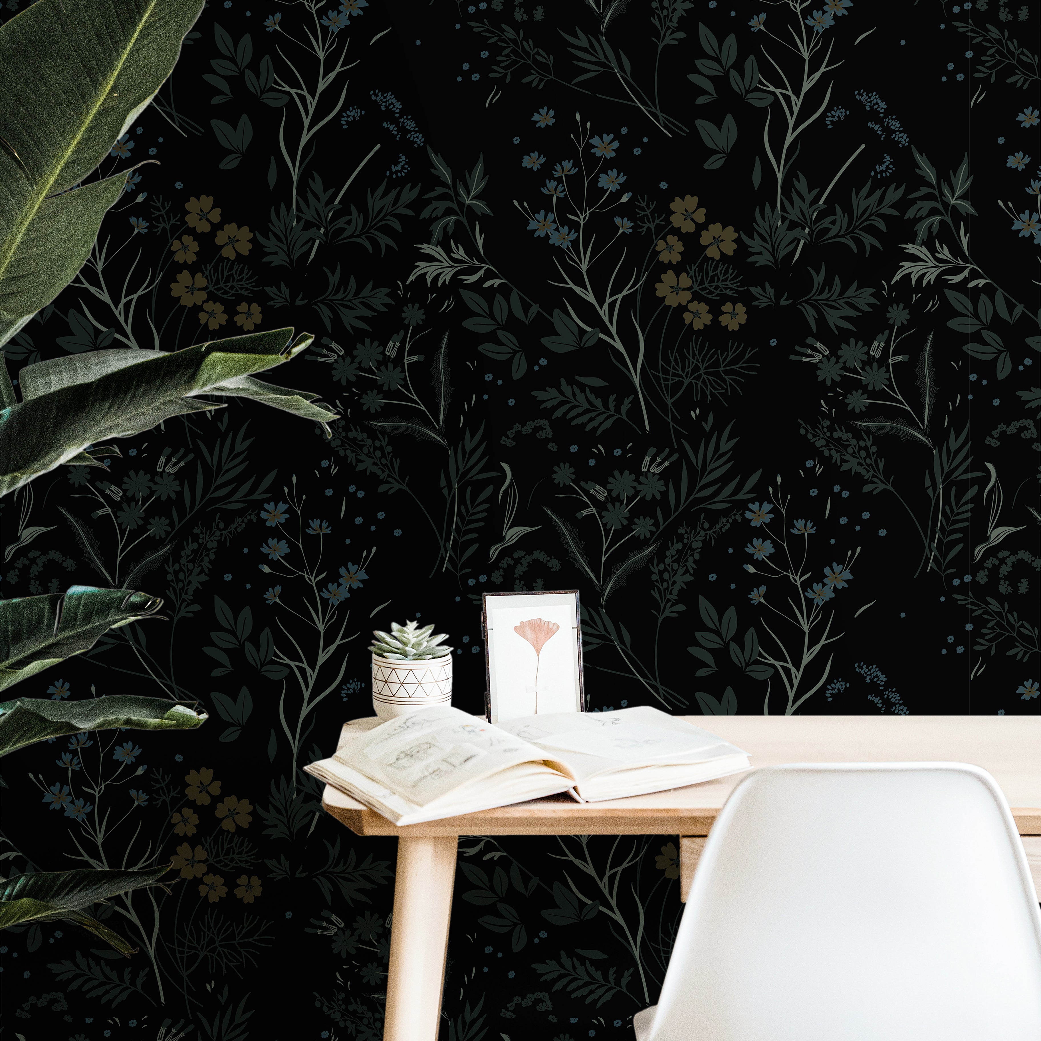 Dark botanical wallpaper featuring layered wildflowers and foliage in muted blue and green tones on a black background styled behind a modern office desk.