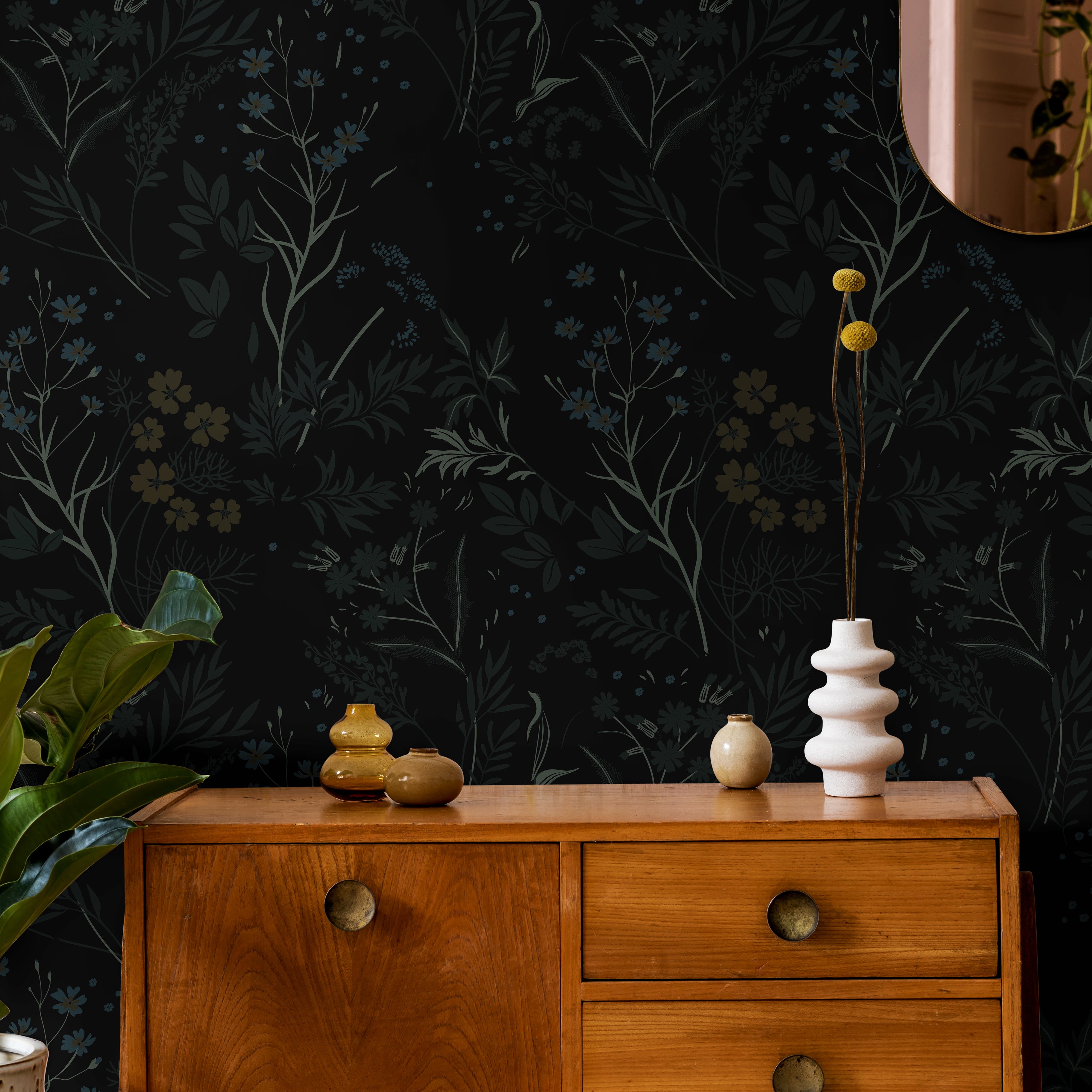 Moody black botanical wallpaper with delicate wildflowers and layered foliage in muted blue, olive, and tan tones styled with warm mid century wood furniture and soft organic decor accents.