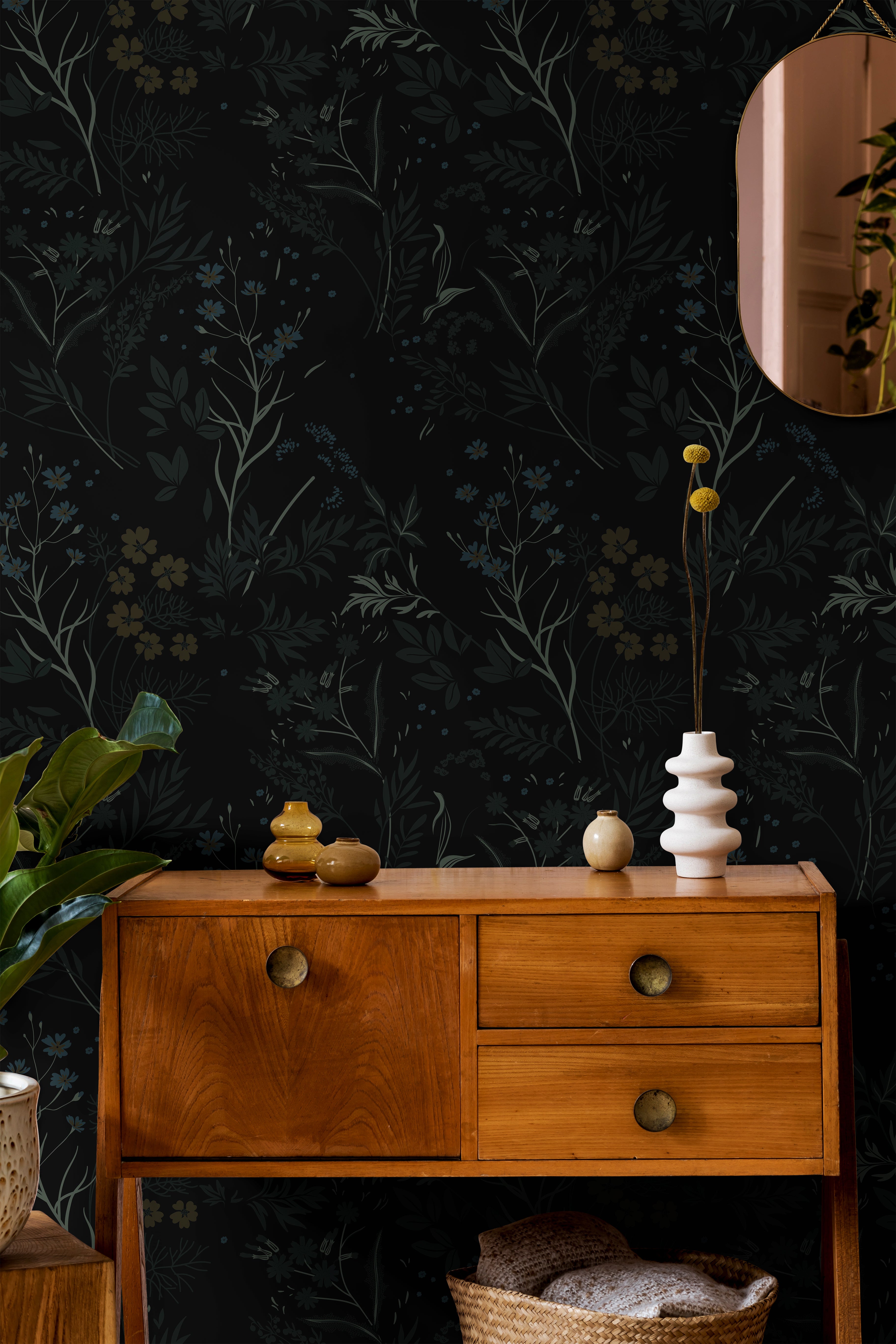 Moody black botanical wallpaper with delicate wildflowers and layered foliage in muted blue, olive, and tan tones styled with warm mid century wood furniture and soft organic decor accents.
