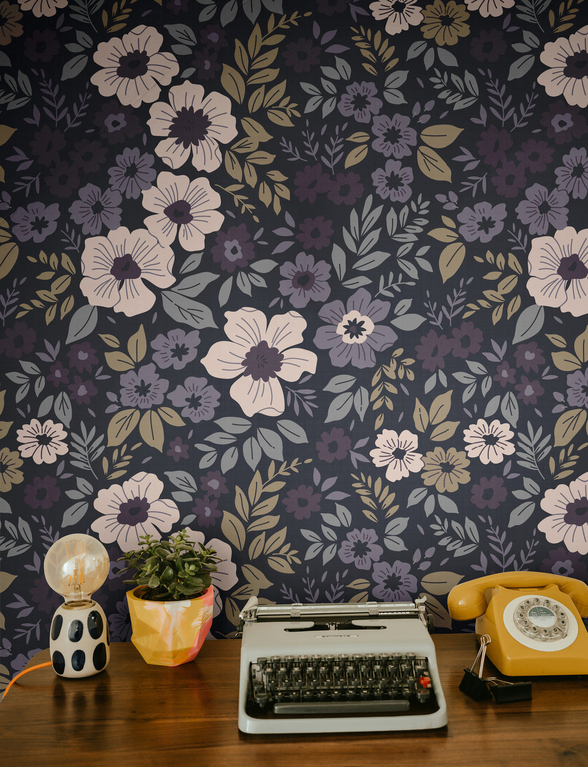 plum and lilac floral wallpaper behind a wood desk with a typewriter yellow phone and potted plant