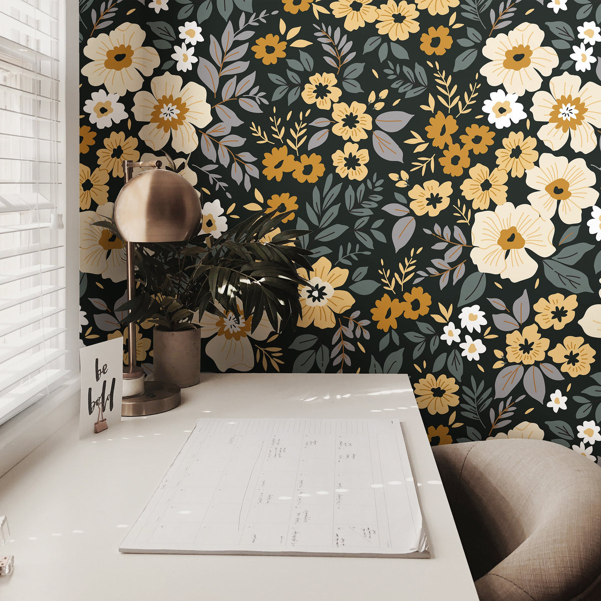 charcoal floral wallpaper behind a sunlit nook with brass lamp potted plant notepad and neutral cushion