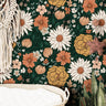 Wallpaper 137: Wallpaper with a bold floral pattern featuring oversized daisies, marigolds, cosmos, and wildflowers in rust, mustard, blush, and cream tones on a deep green background, styled above a rattan changing basket in a boho nursery.

